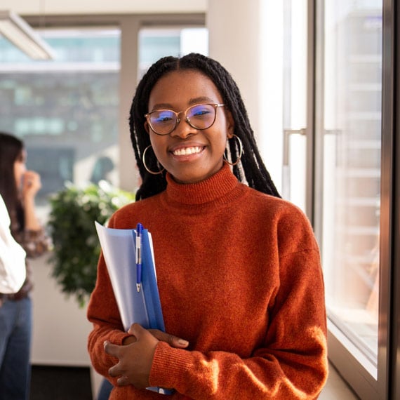 A person wearing an orange sweater holds a blue folder while standing by a window in an office setting