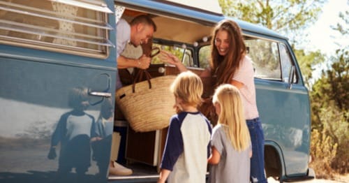 A remote-learning family “roadschooling" with a family on the road getting out of the blue van holding a picnic basket. 