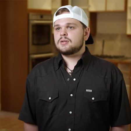Person wearing a black button-up shirt and a white baseball cap in a kitchen.
