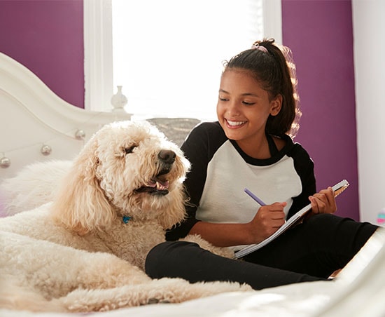 Young girl with her dog sitting on a bed writing in a notebook